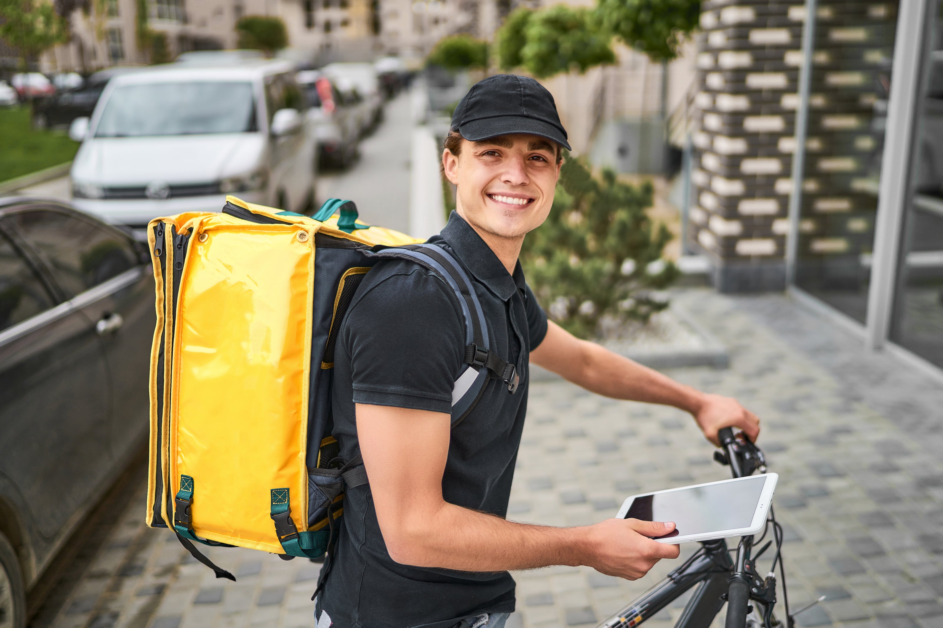 Retrato de feliz repartidor en uniforme en bicicleta cerca de una casa moderna con una mochila amarilla